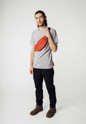 Rote Hüfttasche aus strukturiertem Material, mit einem Reißverschluss und verstellbarem schwarzen Tragegurt, getragen über einem grauen T-Shirt und kombiniert mit schwarzen Hosen.