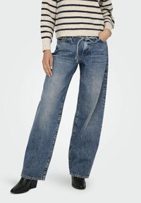 Jeans din denim albastru cu picioare largi, cu o spălare medie, dotate cu o talie cu șnur și buzunare laterale, asortate cu un pulover tricotat cu dungi și cizme negre.