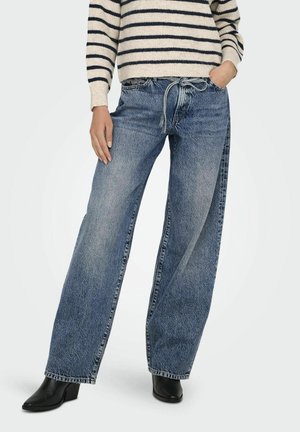 Wijdvallende blauwe denim jeans met een medium wassing, voorzien van een trekkoord in de taille en zijzakken, gecombineerd met een gestreepte gebreide trui en zwarte laarzen.