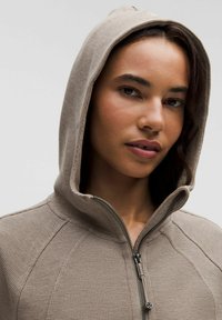 Hanorac crem deschis, cu fermoar, realizat dintr-un material texturat, având glugă și detalii cusute pe cusături, prezentând un design modern, sportiv.