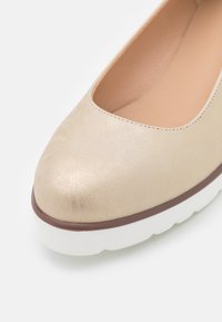 Chaussure beige en cuir à enfiler avec bout arrondi, bordure marron et semelle blanche épaisse texturée.