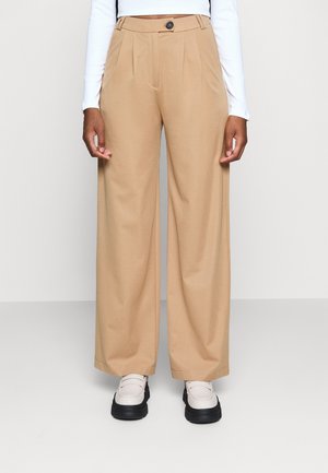 Personne portant un pantalon beige à larges jambes avec bouton à l'avant et plis, un haut blanc à manches longues, et des chaussures blanches à plateforme avec semelles noires.