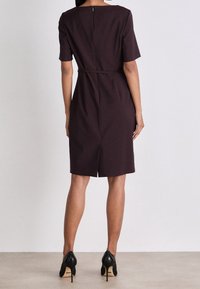 Krátký rukáv, vínová šaty se zipem na zadní straně a páskem v pase, s rovnou siluetou a délkou ke kolenům. Černé lodičky na vysokém podpatku.