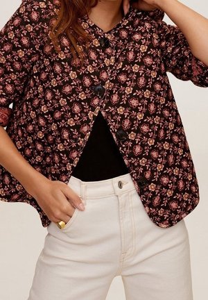 Femme portant une veste boutonnée à motif floral sombre sur un haut noir et un pantalon blanc taille haute, une main dans la poche et l'autre touchant la tête.