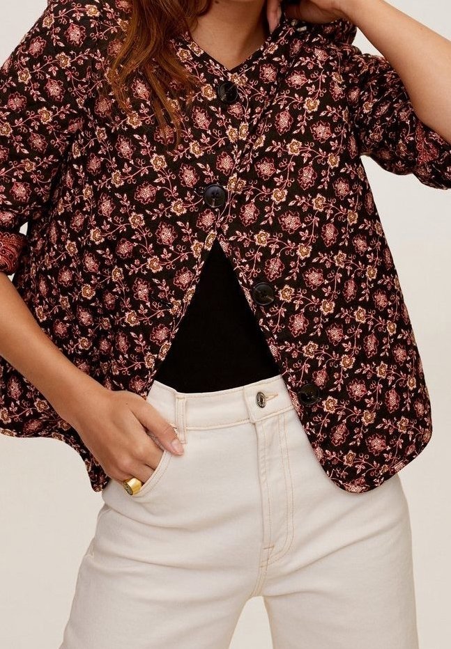 Femme portant une veste boutonnée à motif floral sombre sur un haut noir et un pantalon blanc taille haute, une main dans la poche et l'autre touchant la tête.