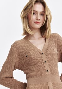 Brązowy dziergany kardigan z dekoltem w kształcie litery V, z teksturowanymi wzorami, guzikami z przodu i dekoracyjnymi metalowymi akcentami przy kieszeniach.