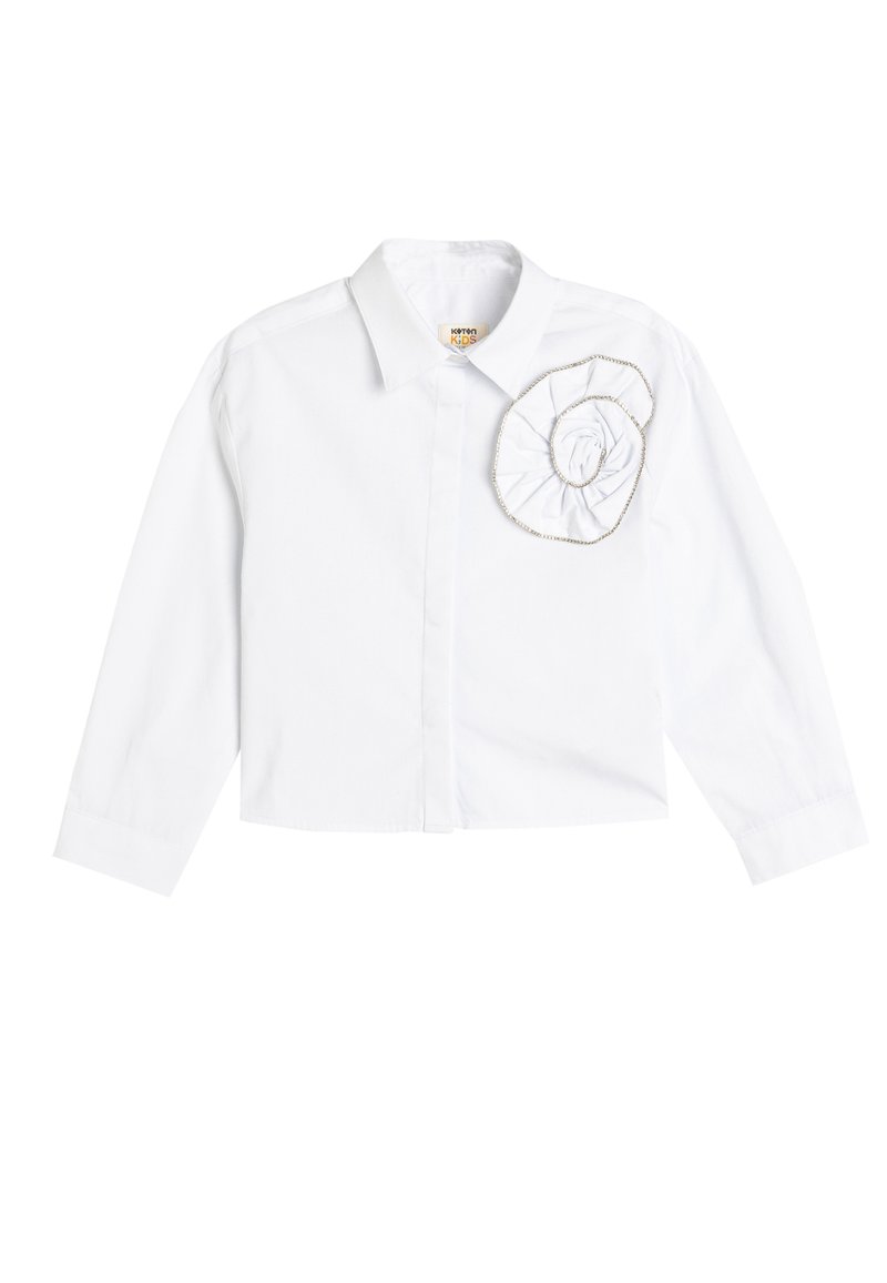 Camisa blanca de manga larga con cuello y adorno de flor de tela en el lado izquierdo del pecho. Hecha de algodón con una textura suave.