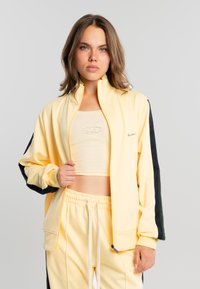 Joven con cabello castaño que lleva una chaqueta y pantalones deportivos amarillo pálido con franjas negras, sujetando la cremallera de la chaqueta.