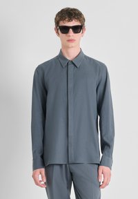Camisa de manga larga y cuello en gris pizarra, fabricada en tejido suave. El modelo lleva gafas de sol oscuras y angulares, de pie frente a un fondo neutral.