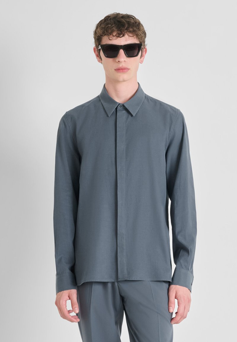 Camisa de manga larga y cuello en gris pizarra, fabricada en tejido suave. El modelo lleva gafas de sol oscuras y angulares, de pie frente a un fondo neutral.