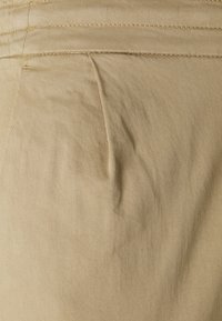 Pantalons en coton beige avec une surface texturée, dotés d'une poche frontale et de coutures visibles. Le tissu présente des plis légers.