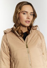 Beige pufferjakke med hætte, med guld lynlås og snore, vandret quiltet mønster og glat tekstur.