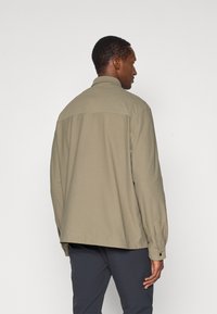 Χακί πουκάμισο με μακριά μανίκια και λεπτομέρεια στο πίσω μέρος, χαλαρή εφαρμογή και κουμπωτές μανσέτες. Κατασκευασμένο από υφαντό ύφασμα με λεία υφή.