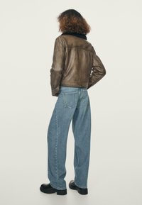 Brązowa skórzana kurtka o skróconym kroju, zestawiona z luźnymi niebieskimi dżinsami i czarnymi masywnymi botkami. Widoczne przeszycia i tylne kieszenie.