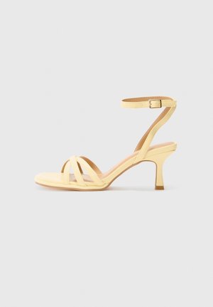 Beige sandal med åben tå med tre stropper foran, ankelrem med spænde og en mellemhøj, slank hæl mod en ensfarvet baggrund.
