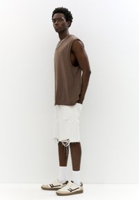 Brown sleeveless top, white distressed shorts, and beige sneakers with dark accents and white socks, worn by a standing model against a white background.
