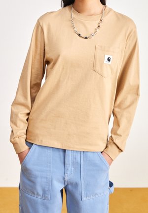 Persoon draagt een beige overhemd met lange mouwen met een logo op het voorzakje en lichtblauwe broek, handen in de zakken en een zilveren ketting om de hals.