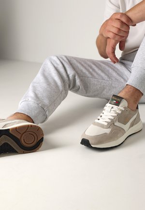 Sneaker beige e bianche realizzate in materiali scamosciati e mesh, con una suola in gomma nera, lacci e una targhetta con logo in accenti verdi e rossi.
