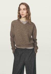 Kobieta z falującymi włosami, ubrana w brązowy sweter w serek na szarym T-shircie i ciemnoszarych szerokich spodniach, z rękami w kieszeniach, na neutralnym tle.