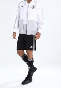 Mužský fotbalista oblečený v bílém a šedém bundě týmu Německa, černých šortkách Adidas, černých ponožkách a černých kopačkách, stál na bílém pozadí.