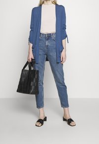 Modrý svetr na knoflíky přes světle růžové tričko, modré džíny s vysokým pasem, černá pletená taška a černé pantofle.