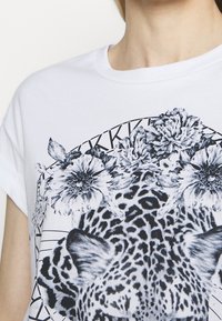 Camiseta de algodón blanca con un gráfico de leopardo en blanco y negro rodeado de elementos florales y patrones geométricos, de corte relajado y mangas cortas.