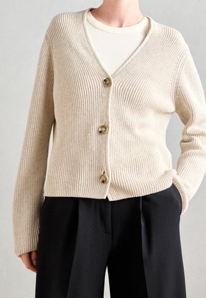 Beige gerippte Strickjacke mit drei Knöpfen über einem weißen Oberteil, kombiniert mit locker sitzender schwarzer Hose, Hand in der Tasche.