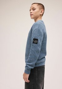 Jasnoniebieski dziany sweter z prążkowanymi mankietami i dołem, z czarną metką na lewym rękawie. Model nosi czarne dżinsy.