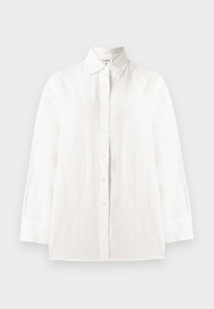 Femme aux longs cheveux foncés portant une chemise blanche boutonnée et un pantalon beige large court, tenant un petit sac tissé rayé.