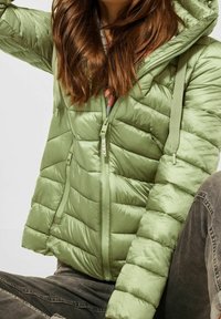 Kvinde iført en lysegrøn quiltet jakke med hætte og lynlås, kombineret med mørkegrå jeans, siddende med korslagte ben.