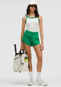 Valkoinen tankkitoppi vihreillä olkaimilla, vihreät shortsit, valkoiset tenniskenkä, ja polvenmittaiset valkoiset sukat. Beigen värinen laukku, jossa on tennismaila ja -pallo.