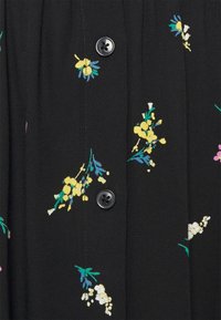Tissu noir avec un tissage texturé, comportant des broderies florales vives en jaune, rose et blanc. Deux boutons noirs à l'avant.