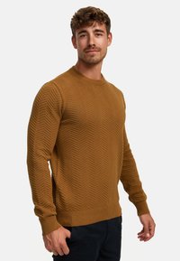 Brown knitted sweater featuring a textured chevron pattern, round neckline, and ribbed cuffs and hem. Worn with dark pants.