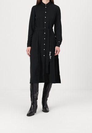 Kobieta w czarnej, długiej, rozpinanej sukience midi z paskiem i czarnych kozakach do kolan, stojąca na białym tle.