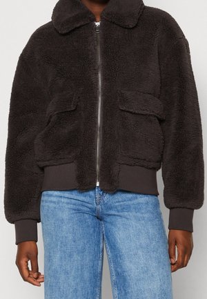 Veste noire duveteuse zippée avec grandes poches avant et poignets côtelés portée avec un jean bleu clair sur une personne à la peau foncée.