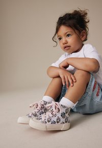 Jeune enfant aux cheveux bouclés assis en tailleur, portant une chemise blanche, un short en jean, des chaussettes blanches et des baskets montantes roses avec des visages de dessins animés.