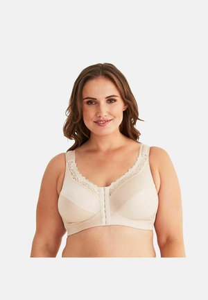 Vrouw glimlacht terwijl ze een beige beha met voorpandsluiting draagt, met kanten bies aan de bandjes en bovenrand, tegen een effen lichte achtergrond.