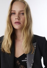 Donna bionda con occhi azzurri indossa un top in pizzo nero e un blazer scuro a righe con ganci d'argento, guardando direttamente la fotocamera su uno sfondo bianco.