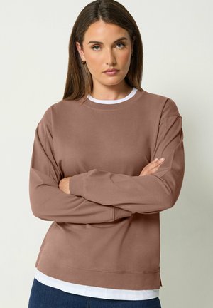 Donna con lunghi capelli castani che indossa una camicia marrone a maniche lunghe sopra una canottiera bianca, in piedi con le braccia incrociate davanti a uno sfondo uniforme.