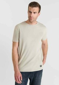 Uomo che indossa una semplice maglietta beige a maniche corte e jeans scuri, in piedi con una mano in tasca e guardando di lato su uno sfondo chiaro.