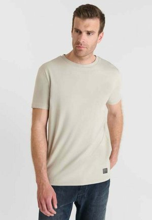 Man in een effen beige shirt met korte mouwen en donkere spijkerbroek, staand met één hand in de zak en kijkend naar de zijkant tegen een lichte achtergrond.