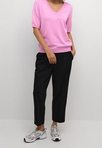 Persona che indossa un top rosa a maniche corte con scollatura a V, pantaloni neri dritti e sneaker bianche su uno sfondo chiaro e uniforme.