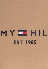 Béžová textilie s žebrovanou strukturou, na které je vyšitý logotyp "TOMMY HILFIGER" a design vlajky v námořnické modré, červené a bílé barvě. Text zní "EST. 1985."