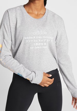 Person iført en lysegrå Adidas sweatshirts med flersproget tekst og sorte Adidas leggings, hånd hvilende nær taljen.