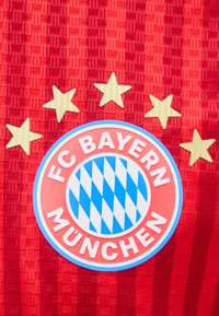 FC Bayern Münchenin logo, jonka yläpuolella viisi kultatähteä punaisessa teksturoidussa urheilupaidan kankaassa.