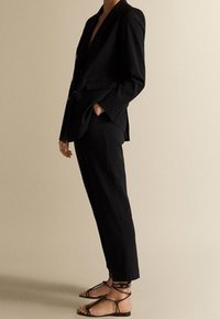 Musta räätälöity puku, jossa on istuva bleiseri ja kapenevat housut. Yhdistetty mustien nilkkaremmillisten sandaalien kanssa. Pehmeä pinta ja siistit linjat.