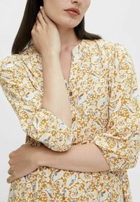 Gelbes Blumenkleid mit Dreiviertelärmeln, das ein Paisleymuster in Weiß, Blau und Braun aufweist. Weiches Material und sichtbare Knopfdetails.