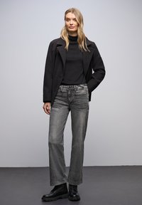 Veste noire, col roulé noir, jeans gris délavé et bottines noires. Silhouette ajustée avec une forme de jambe légèrement décontractée et des détails subtils.