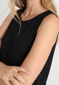 Svart ärmlös topp med en mjuk tygkvalitet, rund halsringning, mjuk drapering och smala armar synliga i bilden, vilket framhäver enkelhet.