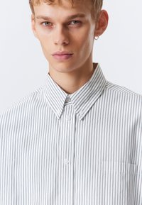 Jovem com cabelo claro, vestindo uma camisa branca de botão com finas riscas verticais pretas e um pequeno brinco em forma de arco na orelha esquerda.